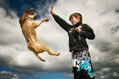 Low angle view of dog fetching stick from boy against cloudy sky