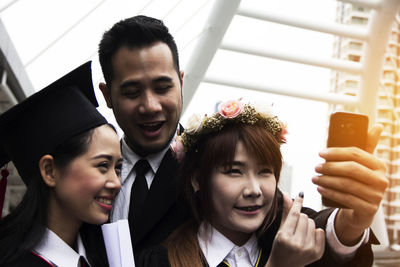 Cheerful young women holding degree certificates taking selfie against sky