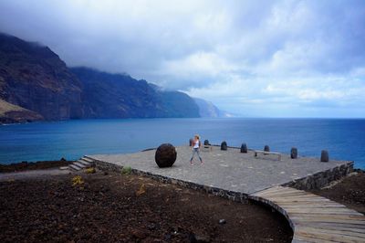 Mid distance view of woman standing at observation point by sea against cloudy sky