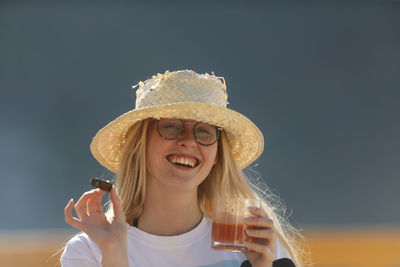 Portrait of smiling young woman wearing hat holding drink and cigar outdoors