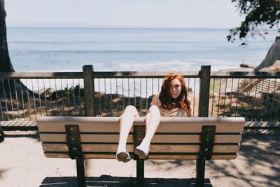 Portrait of young woman sitting on bench by sea