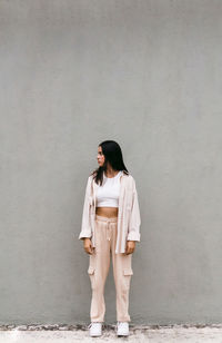 Young female in street style outfit standing on background of gray wall in city and looking away