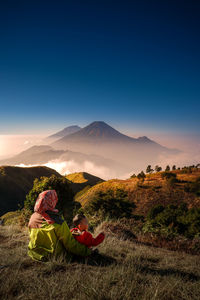 Mother and son sitting on mountain against blue sky