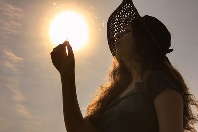 Low angle view of woman blowing dandelion seeds against sun
