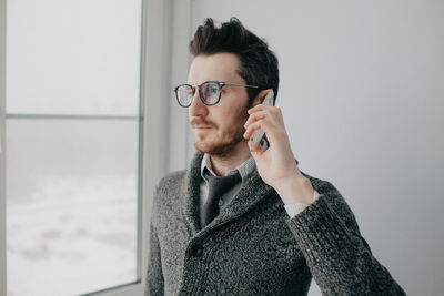 A young stylish man with a beard and glasses in a shirt and a gray cardigan against a gray wall.l