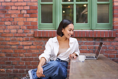 Young man using phone while sitting against brick wall