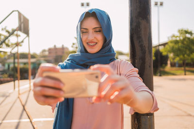 Charming muslim female in traditional headscarf standing in city street and taking self shot on smartphone on sunny day