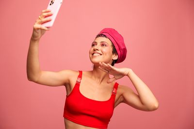 Portrait of smiling young woman taking selfie against pink background