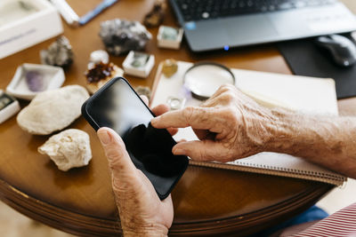 Hands of elderly retired man using smart phone while doing research on fossil and mineral at home