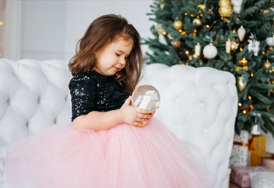 Girl holding showpiece while standing against sofa during christmas