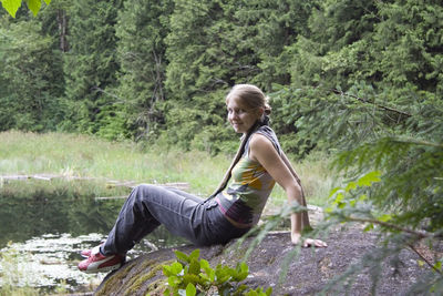 Full length of smiling woman relaxing on rock by pond