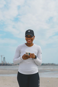 Portrait of young woman holding a smartphone standing smiling at beach against sky