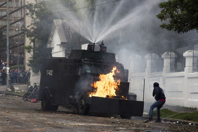 Man standing against burning vehicle spraying water on road in city during protest