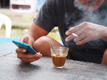 Midsection of man using cigarette having tea while using phone at table