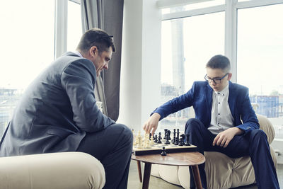 Dad and son are sitting at a table by a wide window and playing chess making moves with pieces