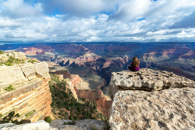 Rear view of woman sitting on cliff against cloudy sky