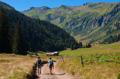 Rear view of hikers walking on dirt road amidst field against mountains