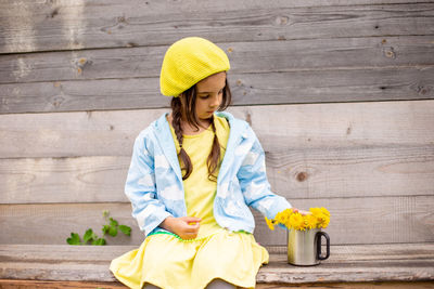  little girl in a yellow beret, blue raincoat with bouquet dandelions in cup near old wooden wall