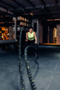 Focused determined young sporty female training with battle ropes during intense workout in modern gym