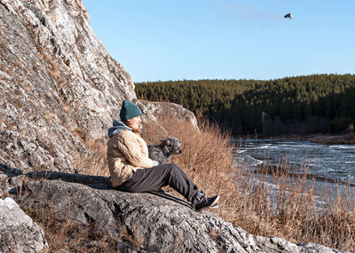 Young woman sitting on stone hugging dog looking at river in first ice in fall or winter beige color