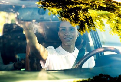 Young woman adjusting rear-view mirror seen through car windshield