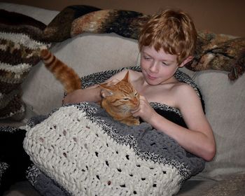 High angle view of shirtless boy playing with cat in knitted blanket