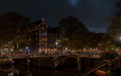 Illuminated bridge over river by buildings against sky at night