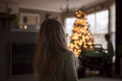 Rear view of sister standing at home while brother by illuminated christmas tree