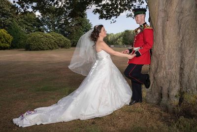 Side view of smiling bride and groom holding hands while standing by tree on field