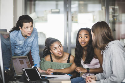 Mature teacher looking at confident female high school students discussing over laptop in computer lab