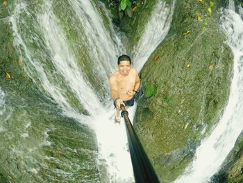 High angle view portrait of shirtless man standing amidst waterfall