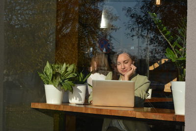 Businesswoman using laptop on table in cafe seen through window
