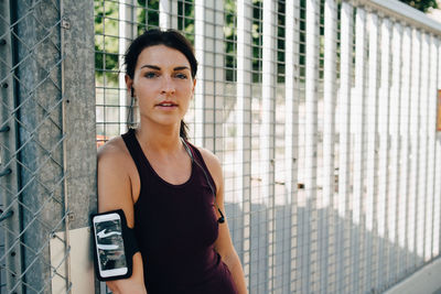 Portrait of confident sportswoman with smart phone and in-ear headphones leaning on railing at bridge