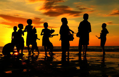 Silhouette boys practicing with musical instruments at beach against cloudy sky during sunset