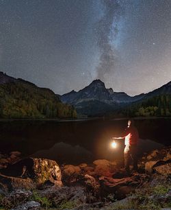 Man standing by lake against mountains at night