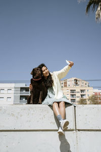 Smiling woman taking selfie with labrador dog while sitting on retaining wall