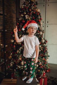 Cute boy stands with christmas tree in red christmas hat, in simple t-shirt, green pajama pants 