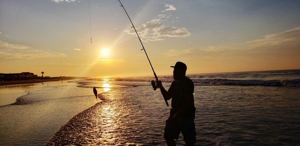 Silhouette man fishing at beach against sky during sunset