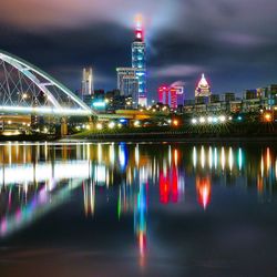 Illuminated modern buildings by river against sky at night