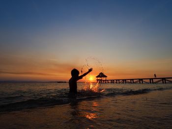 Silhouette boy standing in sea against sky during sunset