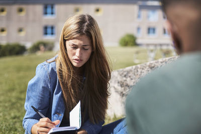 Woman writing on book while sitting with friend at university campus