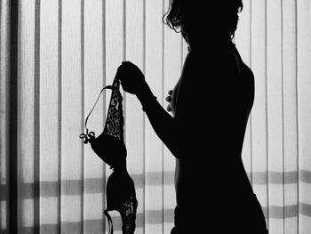 Silhouette sensuous shirtless woman holding bra while standing by curtain at home