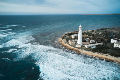 From above drone view of white lighthouse tower and small settlement located on rocky coast washed by foamy sea waves