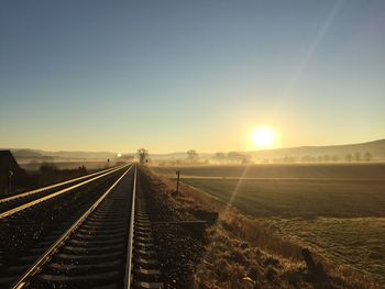 Sunlight streaming on field by railroad tracks against clear sky