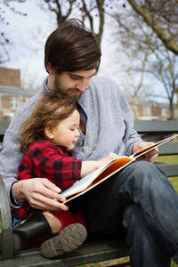 Father and daughter reading book while sitting on bench at park