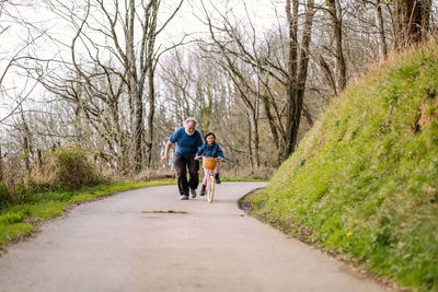 Distant caring grandfather helping active granddaughter riding bicycle with basket on road in countryside with green trees on summer day