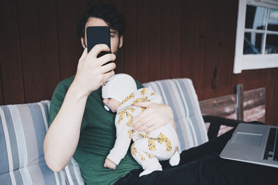 Father with baby boy using mobile phone while sitting on chair against wall