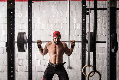 Confident shirtless sportsman standing with heavy barbell and preparing for doing squats during workout in gym