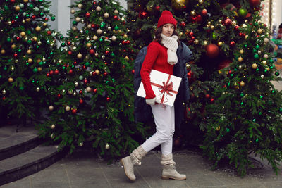 Young woman in a red knitted hat and sweater stands with a gift at the christmas tree