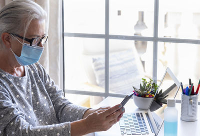 Close-up of senior woman wearing mask using smart phone sitting by window at home